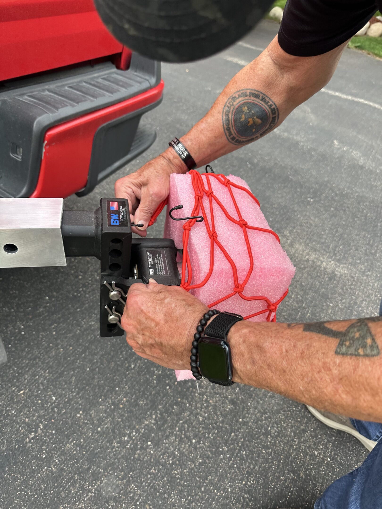 Person securing a red net over a white package on a vehicle hitch.