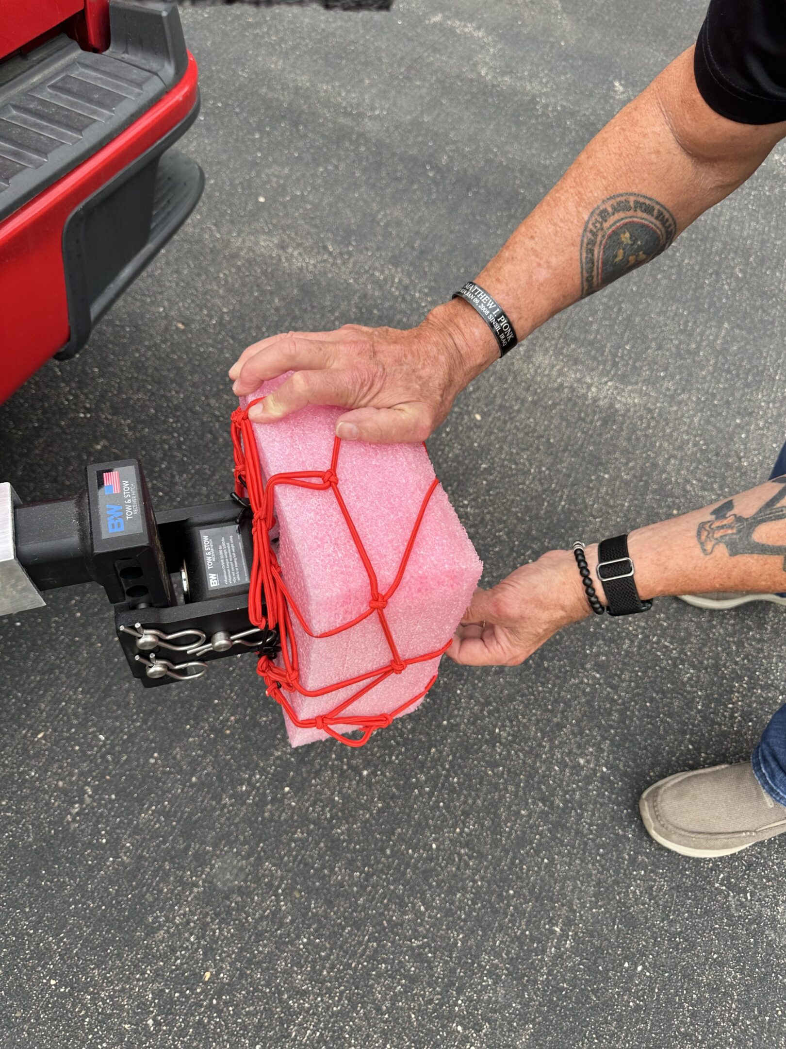 Person securing a red plastic container with a red elastic net on a trailer hitch.